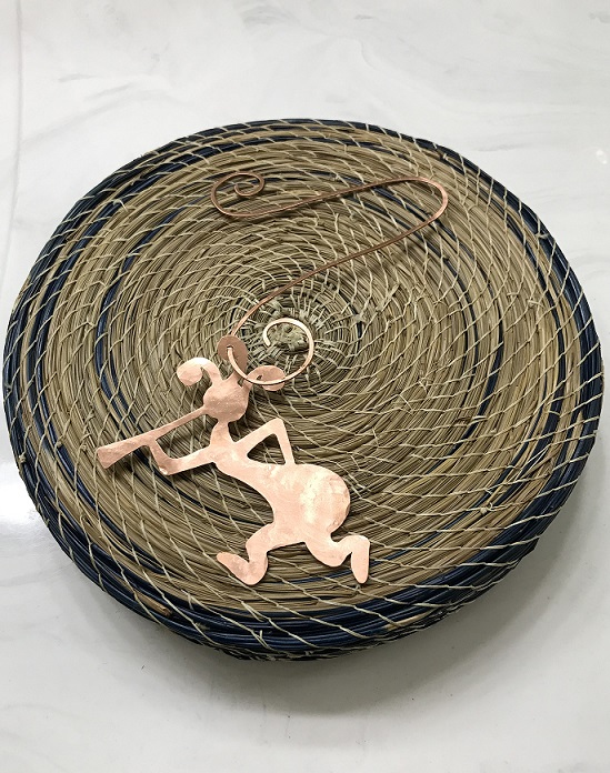 Copper Kokopelli Ornament with Hanger on a blue and tan basket background. This image has more light on the ornament.
